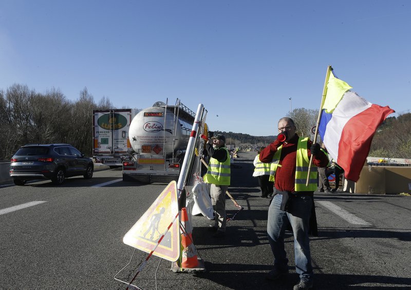 A demonstrator wearing a yellow vest waves the French flag while another one sets up a barrier on a motorway near Aix-en-Provence, southeastern France, Tuesday, Dec. 4, 2018. French Prime Minister Edouard Philippe announced a suspension of fuel tax hikes Tuesday, a major U-turn in an effort to appease a protest movement that has radicalized and plunged Paris into chaos last weekend. (AP Photo/Claude Paris)