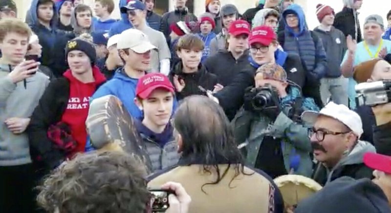 In this Friday, Jan. 18, 2019 image made from video provided by the Survival Media Agency, a teenager wearing a "Make America Great Again" hat, center left, stands in front of an elderly Native American singing and playing a drum in Washington. 