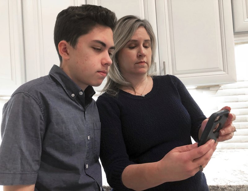 FILE- In this Jan. 31, 2019, file photo Grant Thompson and his mother, Michele, look at an iPhone in the family's kitchen in Tucson, Ariz., on Thursday, Jan. 31, 2019. Apple has released an iPhone update to fix a FaceTime flaw that allowed people to eavesdrop on others while using its group video chat feature. The repair is included in the latest version of Apple's iOS 12 system, which became available to install Thursday. Apple credited the Tucson teenager, Grant Thompson, for discovering the FaceTime bug. (AP Photo/Brian Skoloff, File)