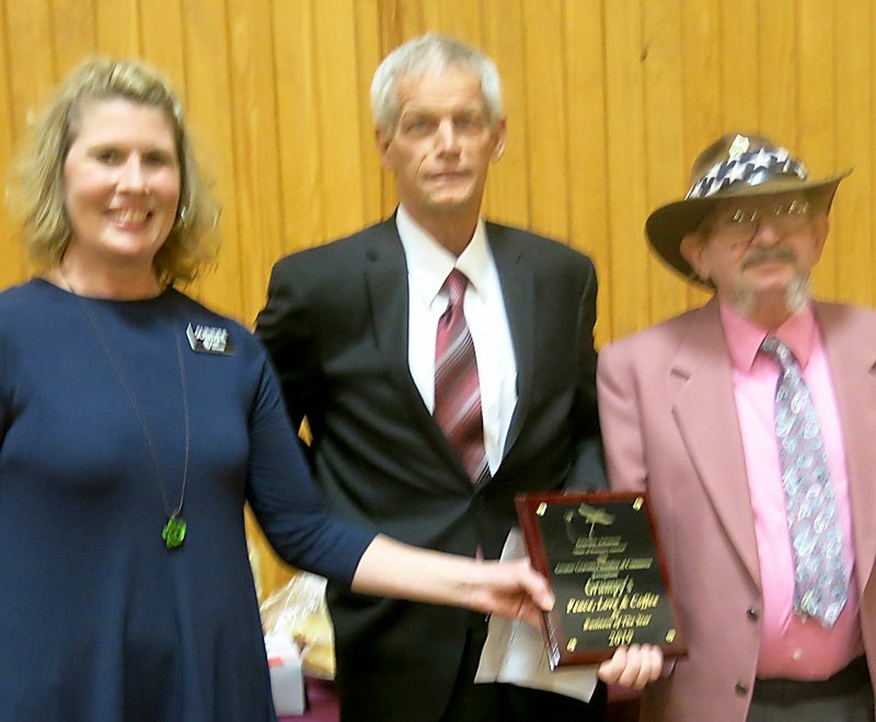 Westside Eagle Observer/SUSAN HOLLAND Gravette Chamber of Commerce treasurer Sundee Hendren and president Fred Overstreet present a plaque to Larry (&#x201c;Grumpy&#x201d;) Jones to denote his business, Grumpy&#x2019;s Peace, Love and Coffee, was chosen Gravette Business of the Year for 2019. Jones was honored at the annual Chamber banquet held Thursday, Feb. 7.