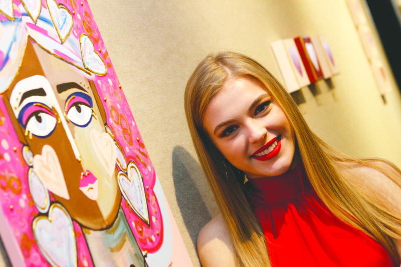 Emmaline Landes, a junior at EHS, poses beside one of her works that is displayed in the lobby of SAAC. Many of her acrylic paintings are available for sale and Landes said the money from selling her artwork goes to different charities that are close to her heart.
