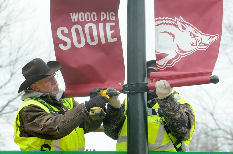 NWA Democrat-Gazette/DAVID GOTTSCHALK Clyde Davis (left) and Earl Perkins, both with University of Arkansas Facilities Management position and install Wednesday, February 20, 2019, banners along Arkansas Avenue on the campus in Fayetteville. The banners are being installed on 58 lamp posts around campus including the Greek Theater area.