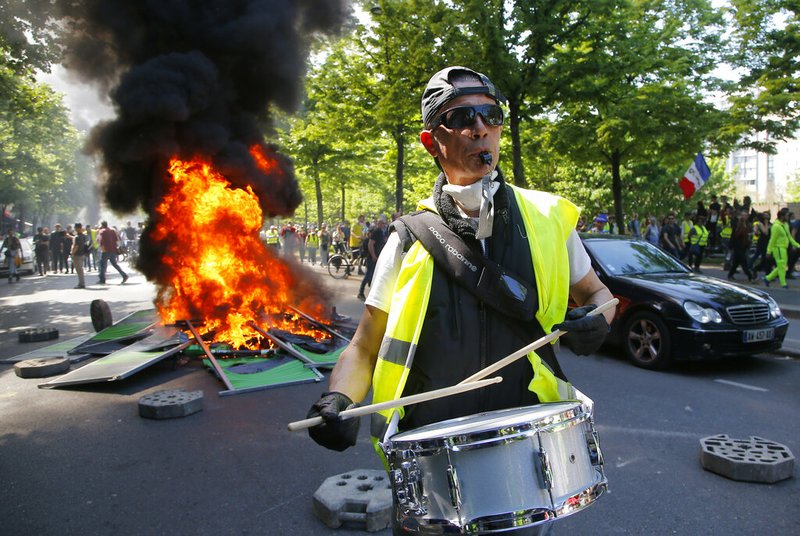 A man bangs a drum in front of a fire on the street during a yellow vest demonstration in Paris, Saturday, April 20, 2019. French yellow vest protesters are marching anew to remind the government that rebuilding the fire-ravaged Notre Dame Cathedral isn't the only problem the nation needs to solve. 