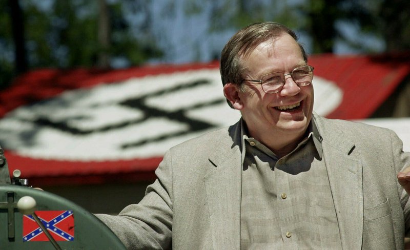 FILE - In this May 22, 2001, file photo, Norm Gissel smiles as he talks about the imminent dismantling of the former headquarters of the Aryan Nations in Hayden Lake, Idaho. Behind Gisel is a Nazi insignia painted atop the roof of the compound's cafeteria. Nearly two decades after the Aryan Nations compound was demolished in Idaho, far-right extremists are maintaining a presence in the Pacific Northwest. (AP Photo/Elaine Thompson, File)