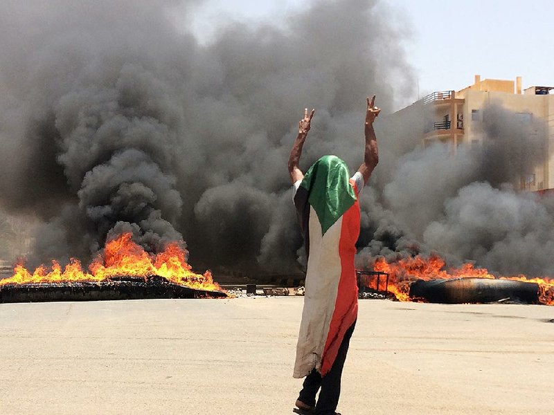 A protester wearing a Sudanese flag flashes the victory sign Monday as tires and debris burn near the military headquarters in Khartoum. 
