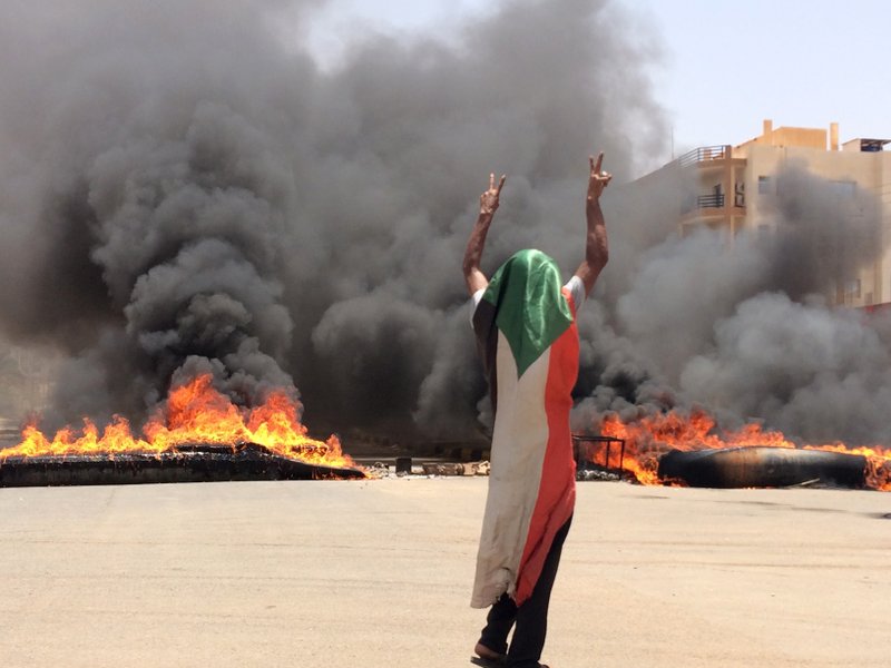 The Associated Press BOMB DEBRIS: A protester wearing a Sudanese flag flashes the victory sign in front of burning tires and debris on road 60, near Khartoum's army headquarters, in Khartoum, Sudan, Monday. Sudanese protest leaders say at least 13 people have been killed Monday in the military's assault on the sit-in outside the military headquarters in the capital, Khartoum. The protesters have announced they are suspending talks with the military regarding the creation of a transitional government.