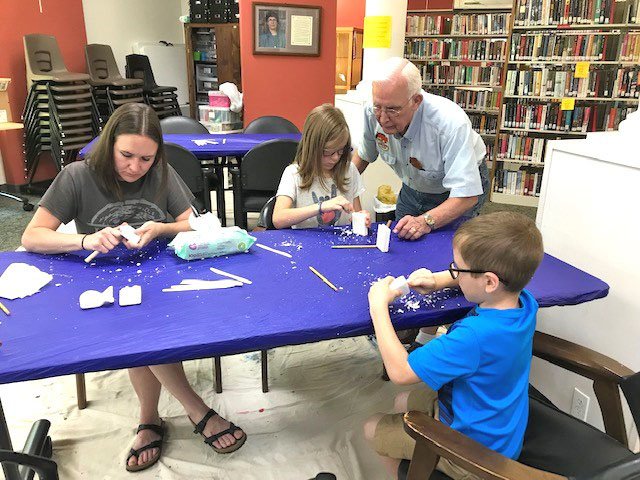 Photo submitted The Bella Vista Woodcarvers Club hosted a soap carving workshop at the Bella Vista Library on June 1. Erin Gandy (left) is shown with her children working on her design. Kinley (top, middle) is shown receiving carving instruction from Lloyd Okeson, and Harper (front, right) is working intently on his project.
