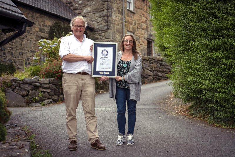 In this undated handout photo provided by Guinness World Records on Tuesday, July 16, 2019, Gwyn Headley and Sarah Badhan, stand on Ffordd Pen Llech with a certificate from Guinness World Records, confirming that the road is the steepest street in the world, in the seaside town of Harlech, North Wales. A street in Wales has been designated the steepest in the world after a successful campaign by local residents. The title comes at the expense of a street in New Zealand which has been eclipsed in the steepness sweepstakes. The Welsh campaign was led by businessman and architectural historian Gwyn Headley. (Andrew Davies/Guinness World Records via AP)