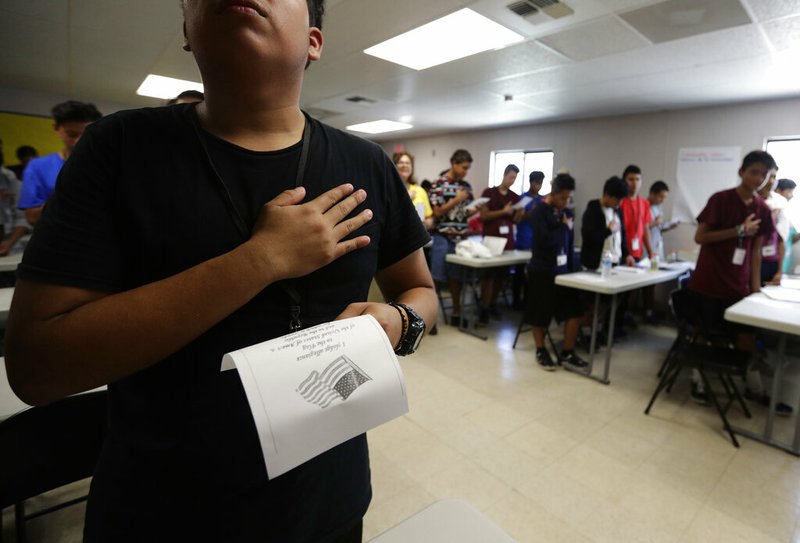 In this Tuesday, July 9, 2019, photo, immigrants say the Pledge of Allegiance in a writing class at the U.S. government's newest holding center for migrant children in Carrizo Springs, Texas. Following breakfast, children play soccer and then have classes held in trailers. (AP Photo/Eric Gay)