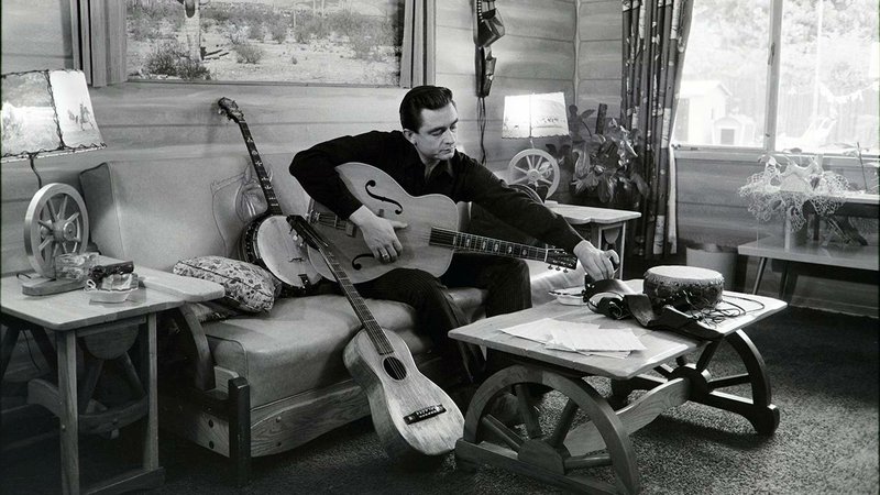 A PBS press photo shows a vintage image of Arkansas native Johnny Cash as he plays a guitar at his home. 