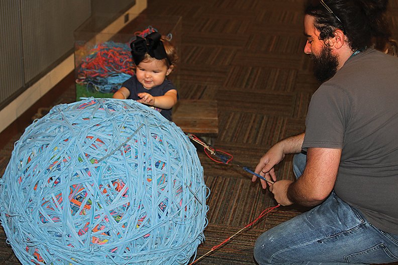 Brooke Hartsfield helps Mid America director of marketing Jim Miller add rubber bands to the over 300 pound rubber band ball on Saturday morning. Next week the ball will crush a car.