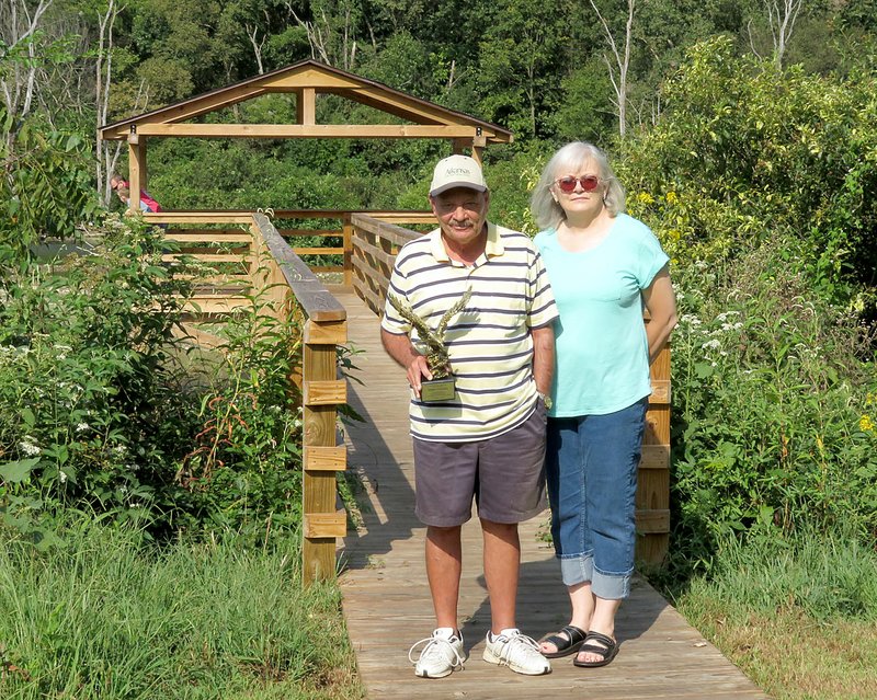 Westside Eagle Observer/RANDY MOLL Terry and Cris Stanfill stand at the entrance to a viewing platform along SWEPCO Lake at the Eagle Watch Nature Area on Saturday (Sept. 14, 2019). Terry Stanfill is holding an award he received for his work to create the nature trail and for his years of service as a manager and promoter of the wildlife area on land owned by the Flint Creek Power Plant. The Stanfills were commended for their contributions to the Eagle Watch Trail at a 20th anniversary celebration Saturday for the nature area.