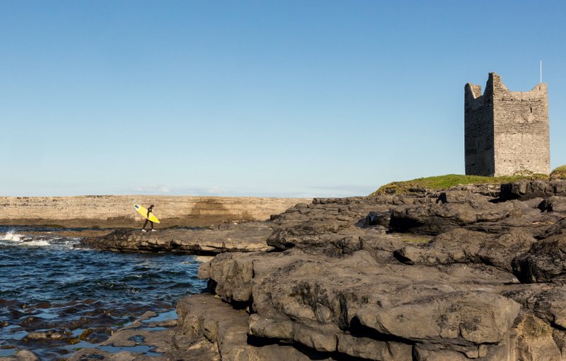 A surfer walks along the shore near the two surf breaks at the mouth of the Easkey River, which flows into the Atlantic Ocean next to a ruin called Roslee Castle in Easkey, Ireland. (Photo by Therese Aherne via The New York Times)