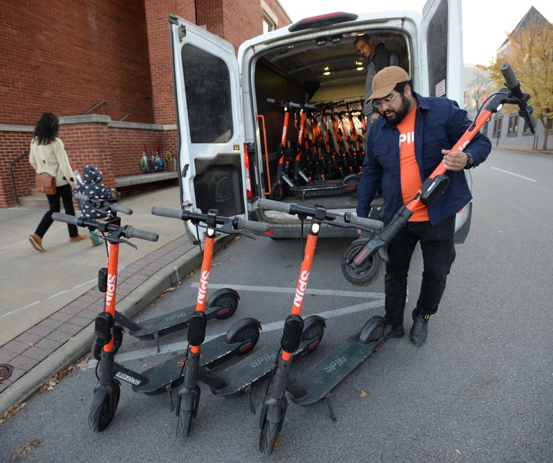Adrian Gomez (right) and Leif Jensen, both operations specialists with Spin unload five of nearly 50 electric scooters near the city administration building in Fayetteville on the first day the scooters will be available for rent in the city with plans to have up to 250 per its contract with the city.. Spin is one of two companies permitted to have electric scooters operating in the city. Users can unlock the scooters with an app on their phones.