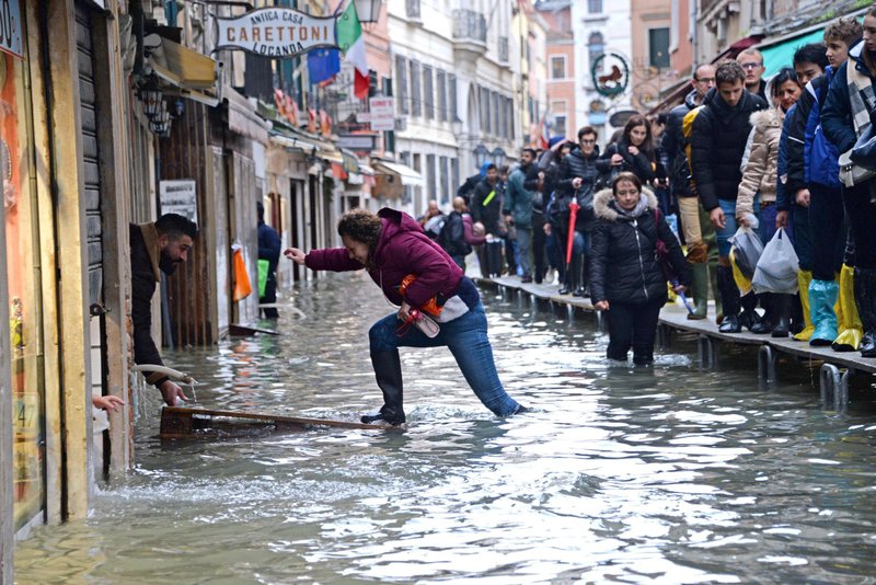 A woman tries to cross a flooded street as people walk on a trestle bridge during high water, in Venice, northern Italy, on Friday. Exceptionally high tidal waters returned to Venice on Friday, prompting the mayor to close the iconic St. Mark's Square and call for donations to repair the Italian lagoon city just three days after it experienced its worst flooding in 50 years. - Andrea Merola/ANSA via AP story.lead_photo.caption