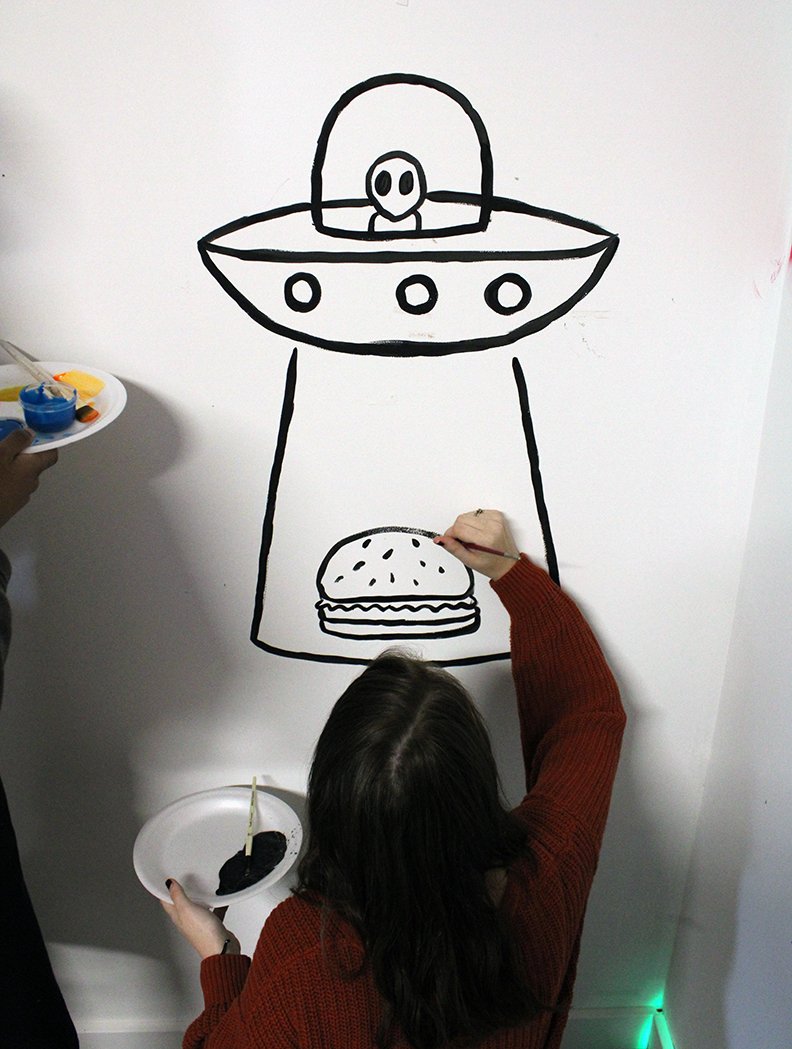 An Upward Bound student, right, paints an alien abducting a hamburger on a wall at the C4K studio. All of the various artworks painted on the walls will be used in a music video that will be shot in the studio in the near future. - Photo by Tanner Newton of The Sentinel-Record