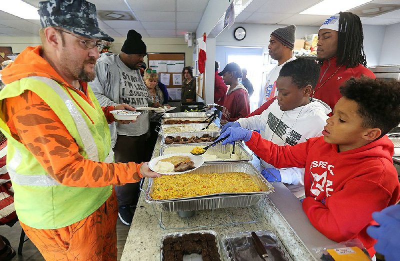 NWA Democrat-Gazette/DAVID GOTTSCHALK Clarke Mitchell (left) receives a spoonful of corn Tuesday, December 31, 2019, from Kingston London, 11, as he volunteers to serve a New Year's Eve meal at the 7 Hills Homeless Center in Fayetteville. The meal was prepared and hosted by Secondhand Smoke NWA and the Northwest Arkansas Generals Youth and Adult Football. The center will be closed today.