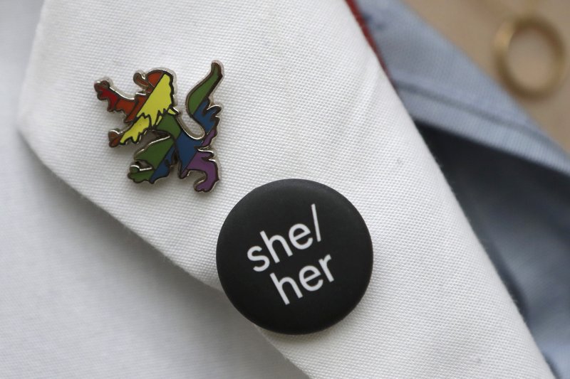 In this Thursday, Oct. 17, 2019 photo Harvard Medical School student Aliya Feroe, of Minneapolis, Minn., displays a button resembling a Harvard School of Medicine coat of arms lion, in rainbow colors that symbolize LGBTQ pride, left, and a button featuring pronouns, center, on the lapel of her lab coat on the school's campus, in Boston. The pronoun button is meant to show support for preferred gender pronouns. Some medical schools are making a big push to recruit LGBTQ medical students, backed by research showing that patients often get better care when treated by doctors who are more like them. (AP Photo/Steven Senne)