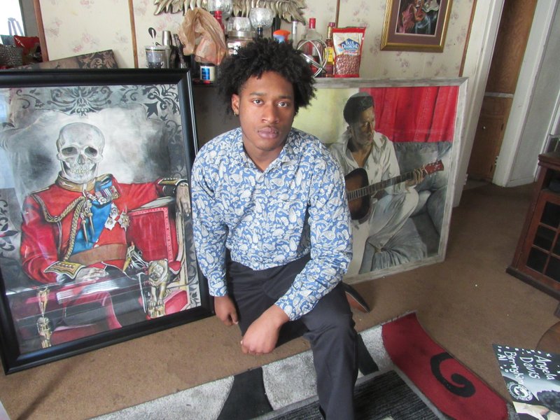 Artist Justin Thomasson poses with two of his most popular works of art at his studio in Pine Bluff. On his right is a self-portrait in which the artist is playing a guitar. On the left is a drawing that, because of the skull and its darker theme, initially alarmed his mother. "I wanted to go for controversy, and this piece is much different from any other piece I created," Thomasson says. (Special to the Democrat-Gazette/Kimberly Dishongh)