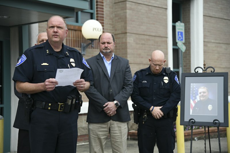 Hot Springs Police Chief Jason Stachey, foreground, reads from a prepared statement at a news conference in front of the Hot Springs Police Department Wednesday evening with, from left, background, Assistant Police Chief Walt Everton, City Manager Bill Burrough, and Cpl. Joey Williams, with a portrait of Officer 1st Class Brent Scrimshire, who was killed in the line of duty on Tuesday. - Photo by Grace Brown of The Sentinel-Record