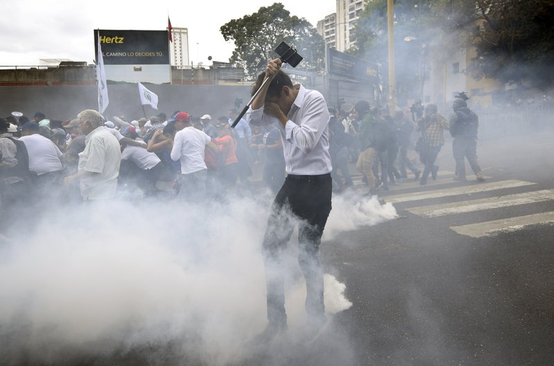 A man holding a cell phone on a selfie stick covers his face amid tear gas fired by police dispersing an opposition march in Caracas, Venezuela, Tuesday, March 10, 2020. U.S.-backed Venezuelan political leader Juan Guaido lead the march aimed at retaking the National Assembly legislative building, which opposition lawmakers have been blocked from entering. (AP Photo/Matias Delacroix)