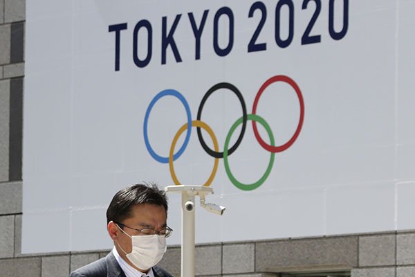 A man walks in front of a Tokyo Olympics logo at the Tokyo metropolitan government headquarters building in Tokyo, Wednesday, March 25, 2020. The Olympic torch relay was postponed Tuesday because the Tokyo Games themselves were pushed back to 2021. (AP Photo/Koji Sasahara)
