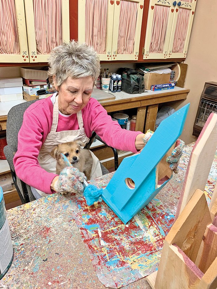Marcia Milligan paints a birdhouse inside the shop she shares with her husband, Gary. Their puppy, Shortcake, finds her usual spot in Marcia’s lap. The Milligans have been crafting together for 40 years.
