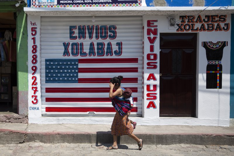 In this April 3, 2020 photo, a woman carrying a child walks past a closed courier business featuring a U.S. flag and the Spanish phrase: "Send to U.S.A" in the largely indigenous town of Joyabaj, Guatemala, where half of the residents depend on remittances, almost all from the U.S. The devastation wrought by COVID-19 across the developed world is cutting into the financial lifelines for people across Latin America, Africa and Asia. (AP Photo/Moises Castillo)
