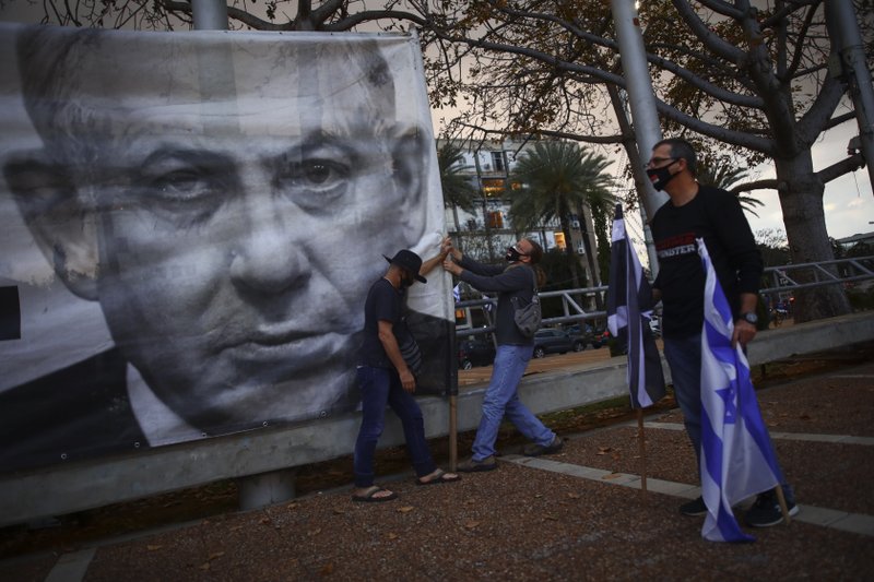 FILE - In this April 25, 2020 file photo, demonstrators wearing protective face masks amid concerns over the country's coronavirus outbreak, hang a banner showing Israeli Prime Minister Benjamin Netanyahu during "Black Flag" protest against Netanyahu and government corruption, at Rabin Square in Tel Aviv, Israel.  (AP Photo/Oded Balilty, File)