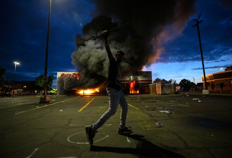 A man poses for a photo in the parking lot of an AutoZone store in flames, while protesters hold a rally for George Floyd in Minneapolis on Wednesday, May 27, 2020.
