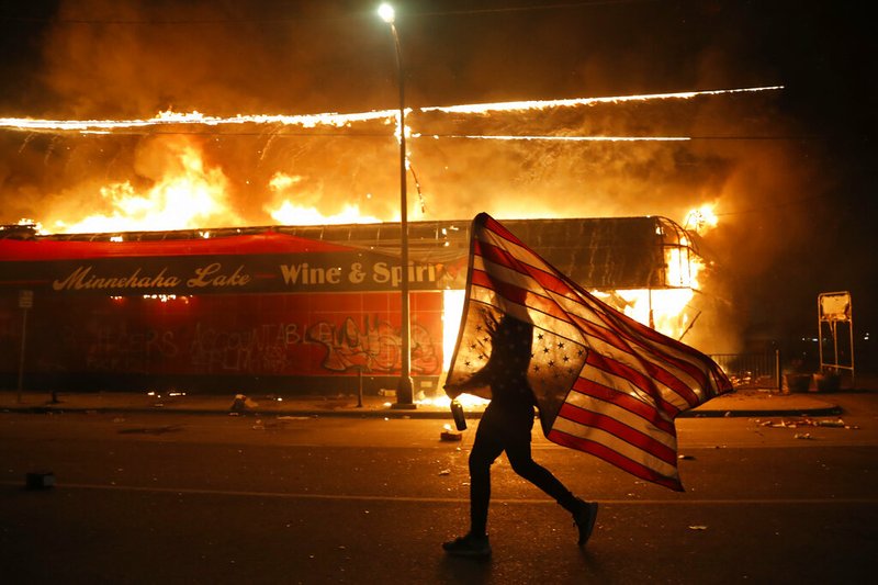 A protester carries a U.S. flag upside, a sign of distress, next to a burning building Thursday, May 28, 2020, in Minneapolis. Protests over the death of George Floyd, a black man who died in police custody Monday, broke out in Minneapolis for a third straight night. (AP Photo/Julio Cortez)

