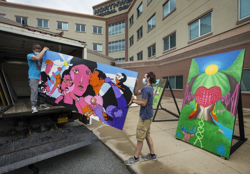 Ian Osgood (left) and Logan Auffet with Lightworks Events in Springdale unload a painting Thursday by artist Octavio Logo at Northwest Medical Center in Bentonville. The men were installing temporary murals by nine local artists that are part of the Social Connecting Campaign by Crystal Bridges Museum of American Art and the Momentary. The campaign seeks to support those most vulnerable to the effects of isolation during the covid-19 pandemic. People have been taking home postcards with line art versions of the paintings to color and mail to hospital patients and senior living facilities. The murals themselves are on a tour of the same facilities throughout Northwest Arkansas, set up each day outside for residents and guests to enjoy. (NWA Democrat-Gazette/Ben Goff)