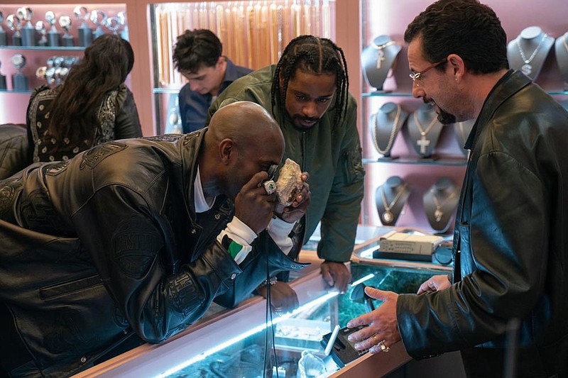 Basketball star Kevin Garnett (left, playing himself), having been steered to Howard Ratner’s (Adam Sandler, right) diamond district jeweler shop by the fixer Demany (LaKeith Stanfield), examines a raw opal that might have mystical powers in the Safdie brothers’ Uncut Gems.