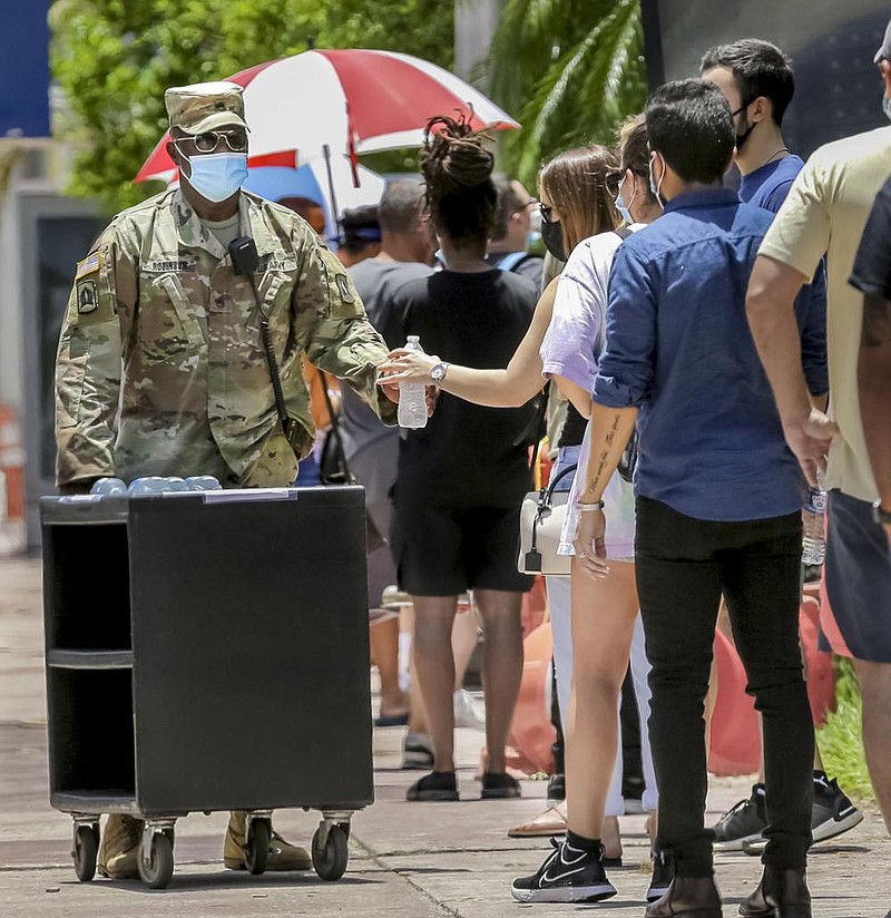 A member of the Florida National Guard passes out bottled water to people waiting in line Tuesday at a walk-up coronavirus testing site in Miami Beach. Florida reported 152,434 covid-19 cases Tuesday, up 4.2% from a day earlier. More photos at arkansasonline.com/71virus/. (AP/Lynne Sladky) 