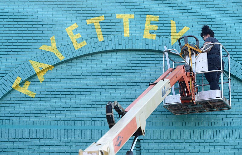 Artist Olivia Trimble, owner of Sleet City Signs and Murals, moves a lift back into position Wednesday, August 12, 2020, to continue painting a new mural on the backside of the Experience Fayetteville building on the square in downtown Fayetteville. The mural is yet to be named and the finished words remain a surprise until the mural is complete. (NWA Democrat-Gazette/David Gottschalk)