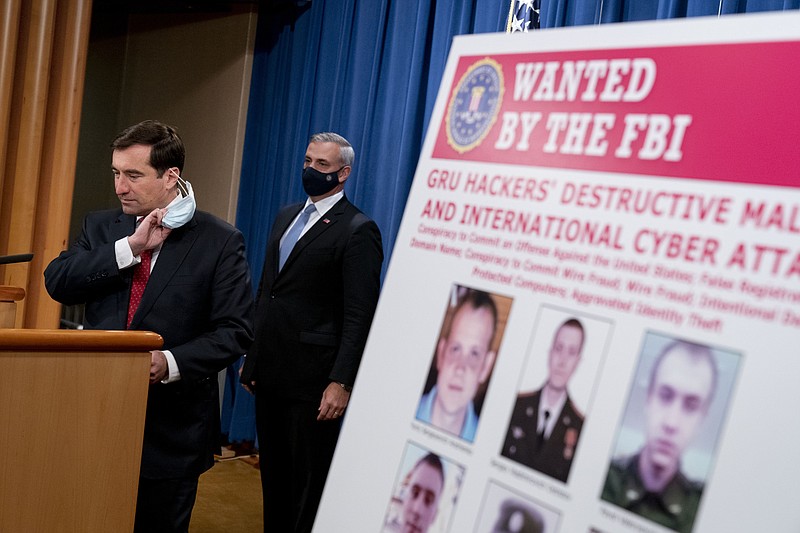 A poster showing six wanted Russian military intelligence officers is displayed as Assistant Attorney General for the National Security Division John Demers, left, takes the podium to speak at a news conference at the Department of Justice, Monday, Oct. 19, 2020, in Washington. Also pictured is US Attorney for the Western District of Pennsylvania Scott Brady, center.