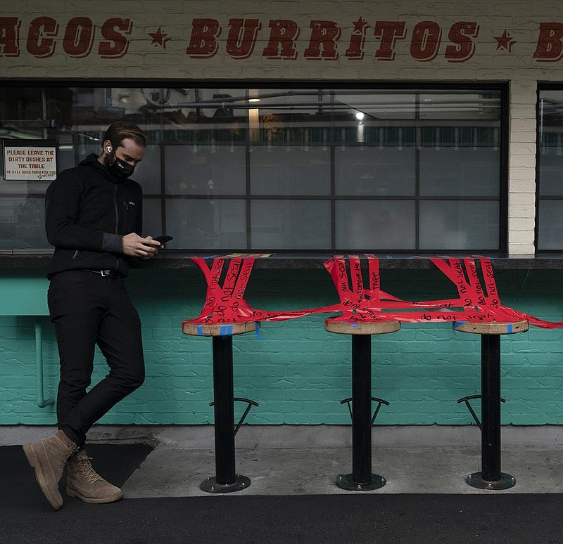 A man looks at his phone next to stools taped off to prevent diners from sitting on them in Los Angeles, Monday, Dec. 7, 2020. The vast region of Southern California went into a lockdown Monday in an effort to curb spiraling coronavirus infections and hospitalizations. (AP Photo/Jae C. Hong)