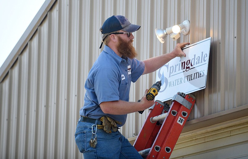 Andrew Brown, pump station operator, hangs a new sign at the former Bethel Heights wastewater treatment facility in Springdale in this Aug. 24, 2020, file photo. The sign signifying the building as a part of Springdale's utility system came less than two weeks after the Benton County Election Commission certified the results of an election to annex Bethel Heights into Springdale because of the town's failing sewer system. (NWA Democrat-Gazette/David Gottschalk)