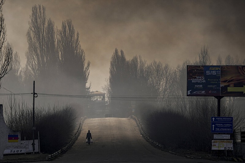 A man carries shopping bags as heavy smoke from a warehouse destroyed by Russian bombardment casts shadows on the road outside Kyiv, Ukraine, Thursday, March 24, 2022. (AP/Vadim Ghirda)