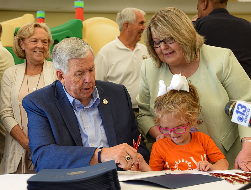 Debbie Hamler, background left, the longtime executive director of The Special Learning Center, looks on and smiles as Missouri Gov. Mike Parson, seated at left, helps six-year-old Abby Fisher hold the paper down so she could sign her name to HB 3020 during a signing ceremony Wednesday, July 20, 2022, at SLC. Standing in the background middle is Rep. Bruce Sassman, R-Bland, who supported this legislation through the House of Representatives. Standing at right is Stephanie Johnson, current executive director at SLC. Fisher, who will attend kindergarten this fall, arrived at SLC at a very young age in a wheelchair, and with therapy and hard work moved up to a walker and later crutches, and now can walk independently on her own. (Julie Smith/News Tribune photo)