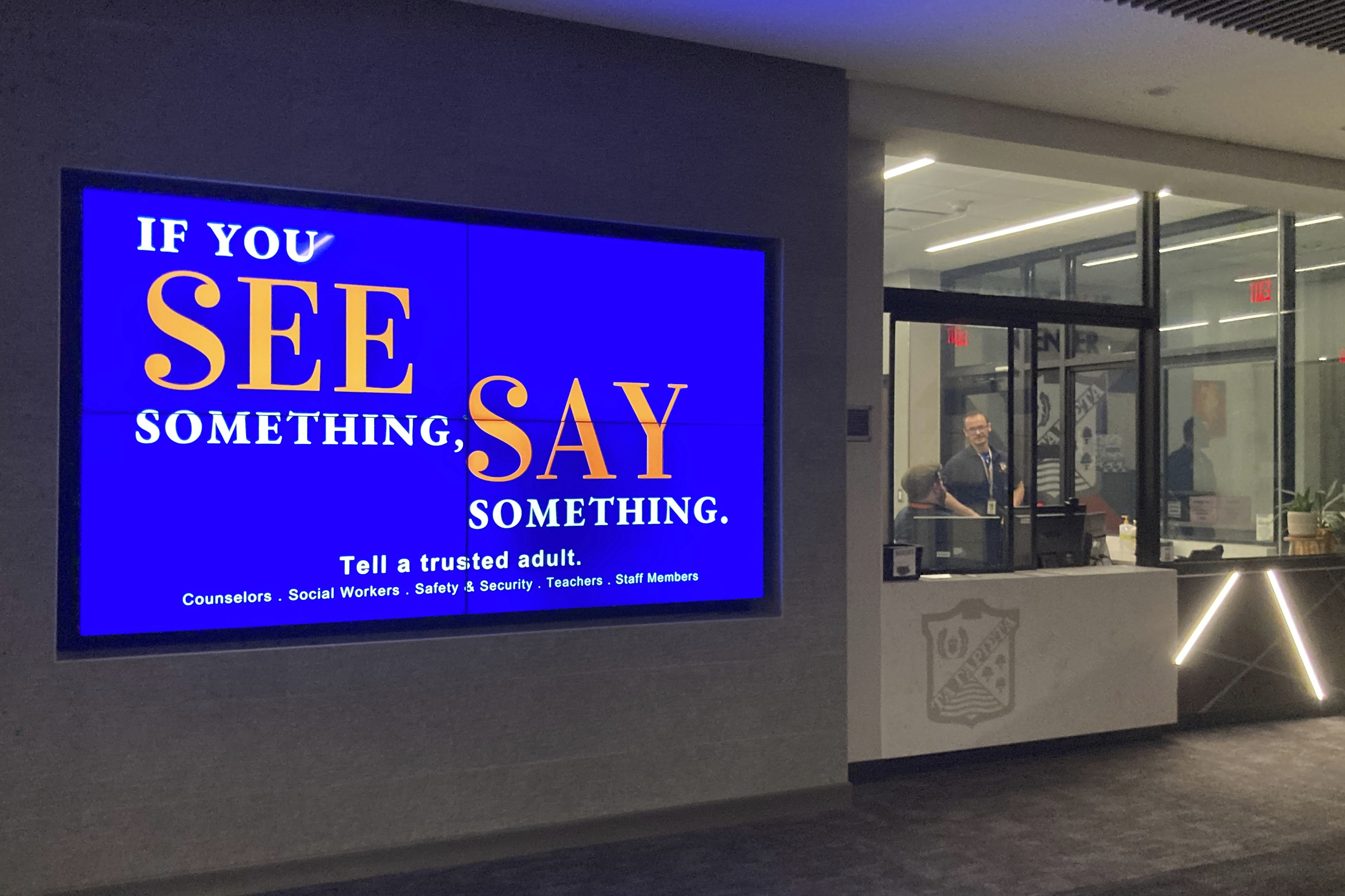 With "See Something, Say Something" displayed on a main entrance monitor in the foreground, security staff chat at Oak Park and River Forest High School after a school board meeting on Thursday, Aug. 11, 2022, in Oak Park, Ill. Signs encouraging students to report suspicious and unsafe activity can be found throughout this and many other high schools. (AP Photo/Martha Irvine)