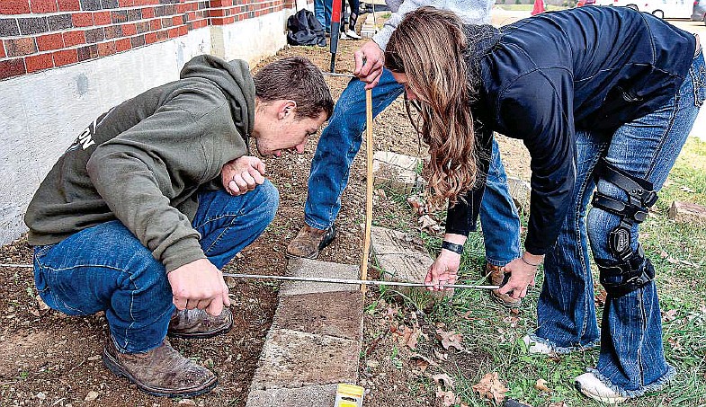 Taylor Brand and Dorothy Lackman measure the distance from the wall to the front of the block Monday, Nov. 14, 2022, while installing a border as part of a class project. Students taking a landscaping segment at Blair Oaks High School have been working at the elementary school to construct a block border on the building's main entry side. (Julie Smith/News Tribune photo)