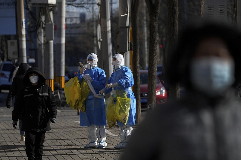 People walk by workers in protective suits looking for the location of lockdown residents to collect covid-19 samples Thursday in Beijing. More photos at arkansasonline.com/122cncovid/.
(AP/Andy Wong)