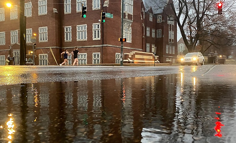 Staff Photo by Robin Rudd / The University of Tennessee at Chattanooga's Gary W. Rollins College of Business is reflected in a McCallie Avenue puddle as two runners cross Douglas Street on the morning of Dec. 6, 2022.