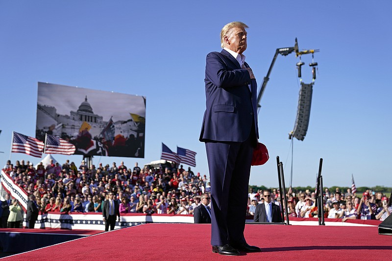 As footage from the Jan. 6, 2021, insurrection at the U.S. Capitol is displayed in the background, former President Donald Trump stands while a song, "Justice for All," is played during a campaign rally at Waco Regional Airport, Saturday, March 25, 2023, in Waco, Texas. The song features a choir of men imprisoned for their role in the Jan. 6, 2021, insurrection singing the national anthem and a recording of Trump reciting the Pledge of Allegiance. (AP Photo/Evan Vucci)