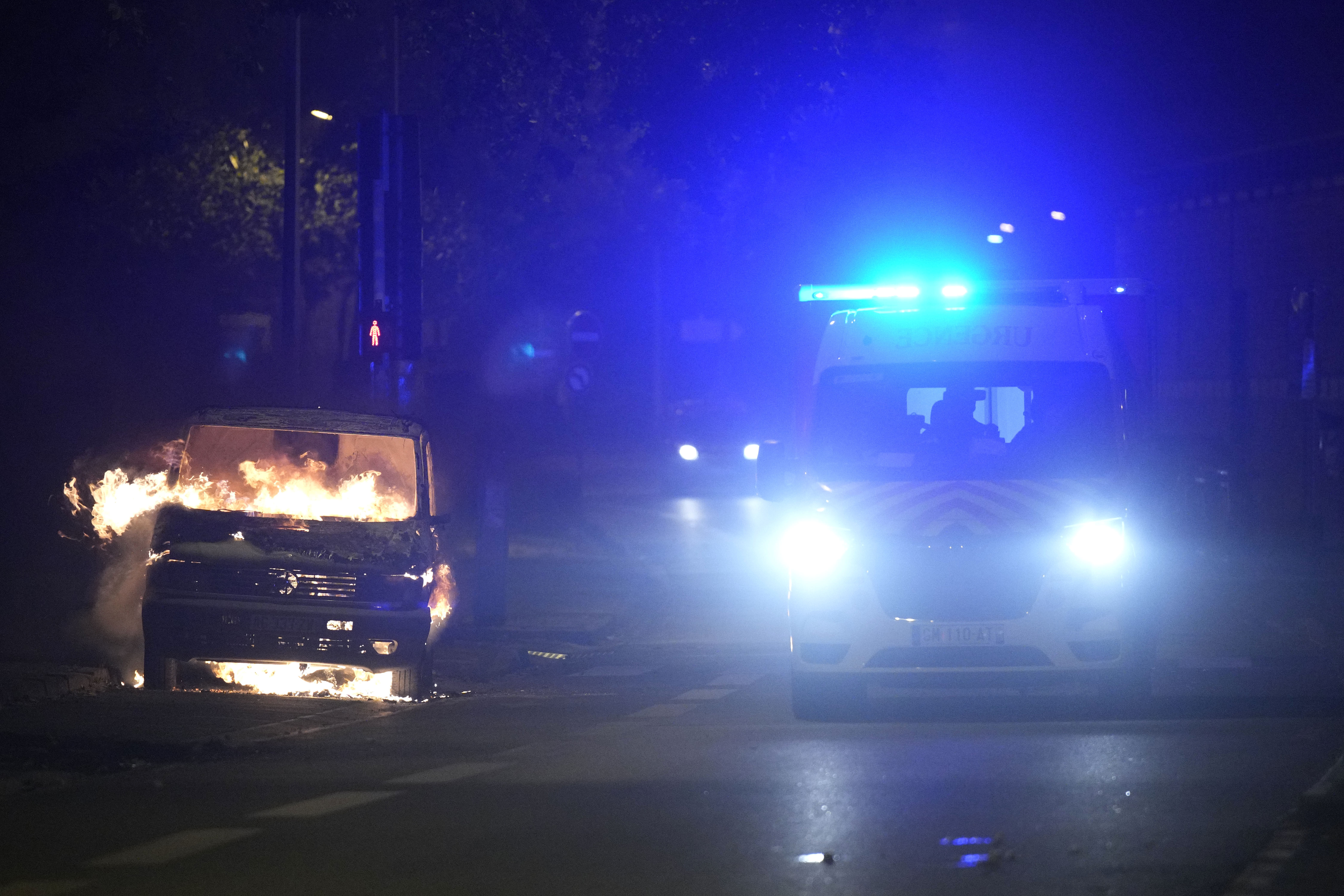 An ambulance passes by burning car in Nanterre, outside Paris, France, Saturday, July 1, 2023. French President Emmanuel Macron urged parents Friday to keep teenagers at home and proposed restrictions on social media to quell rioting spreading across France over the fatal police shooting of a 17-year-old driver. (AP Photo/Lewis Joly)