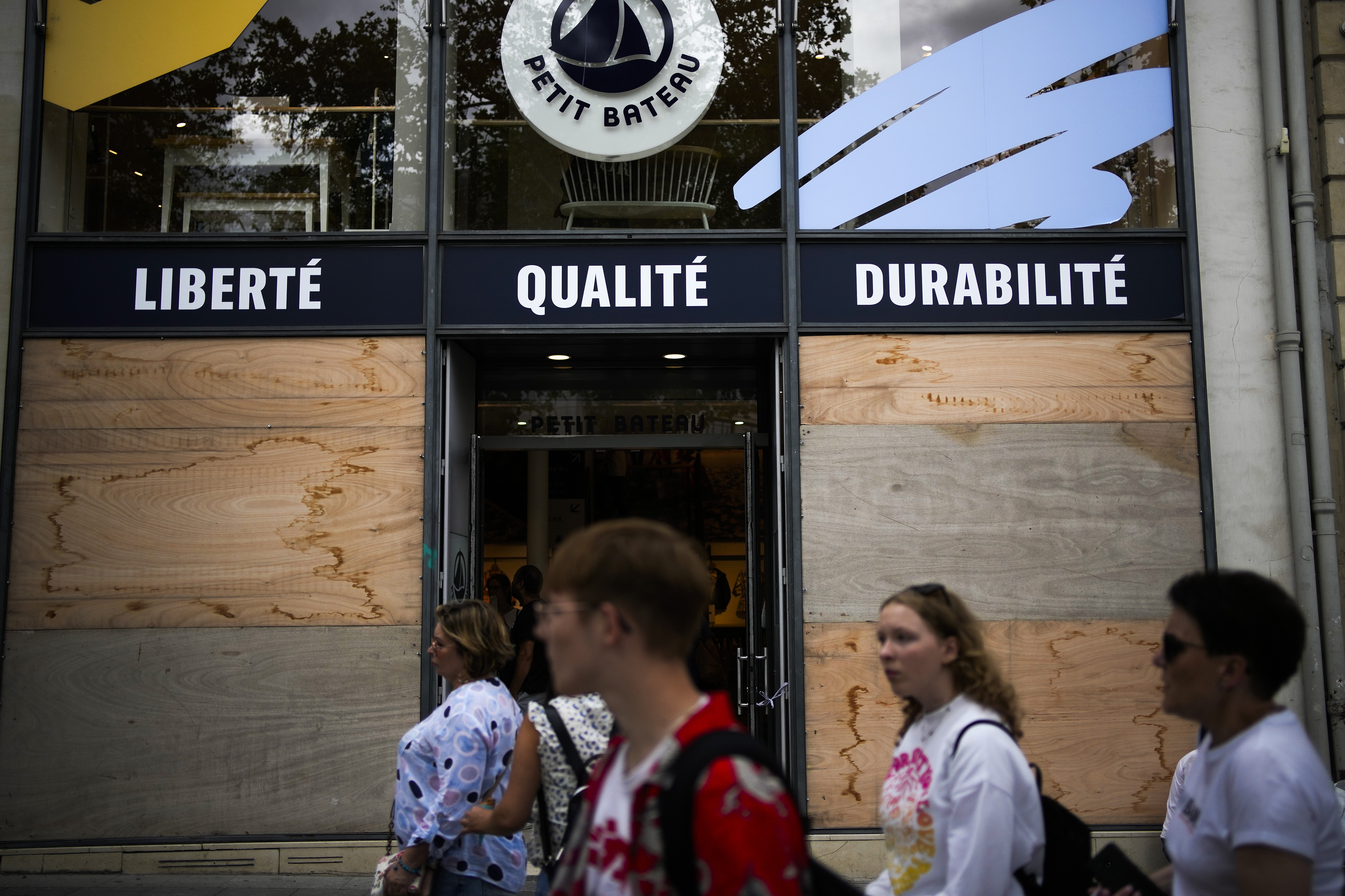 People walk past a shop covered with protection wood planks on the Champs-Elysees avenue in Paris, Saturday, July 1, 2023. The fatal shooting of Nahel, whose last name has not been made public, stirred up long-simmering tensions between police and young people in housing projects who struggle with poverty, unemployment and racial discrimination. (AP Photo/Christophe Ena)