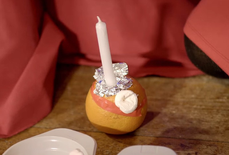 A Christingle — an orange with a candle placed in the top — is shown in this undated screenshot from video taken at All Saints High Wycombe in Oxford, England. Visible on the Christingle is a red ribbon, representing the love of Christ, and a piece of candy attached with a toothpick. (Photo courtesy the Church of England, via YouTube)