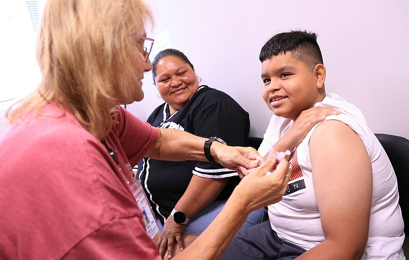 Cristian Maradiaga (10), right, receives a COVID-19 vaccination from Arkansas Department of Health nurse Rita Wilson, left, as his mother Wendy Rios looks on during a vaccine clinic hosted by the Little Rock School District at the Pulaski County Health Department on Tuesday, July 23, 2024. (Arkansas Democrat-Gazette/Colin Murphey)