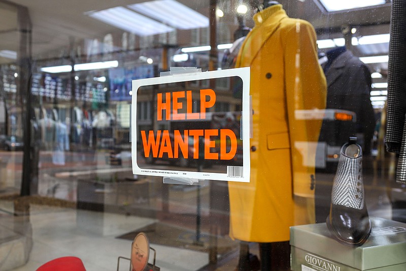 Staff Photo by Olivia Ross  / A "help wanted" sign is seen posted in the window of Giorgio Men's Warehouse as a U.S. Postal Service worker walks in with packages on Tuesday.